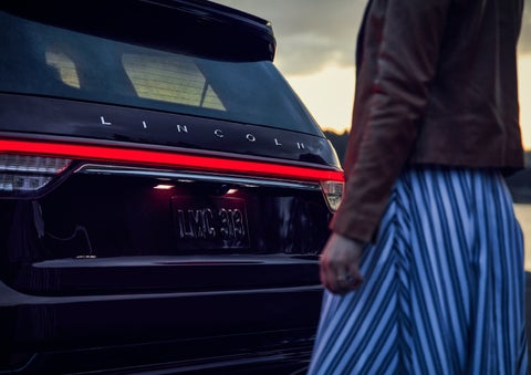 A person is shown near the rear of a 2024 Lincoln Aviator® SUV as the Lincoln Embrace illuminates the rear lights | Performance Lincoln Bountiful in Bountiful UT